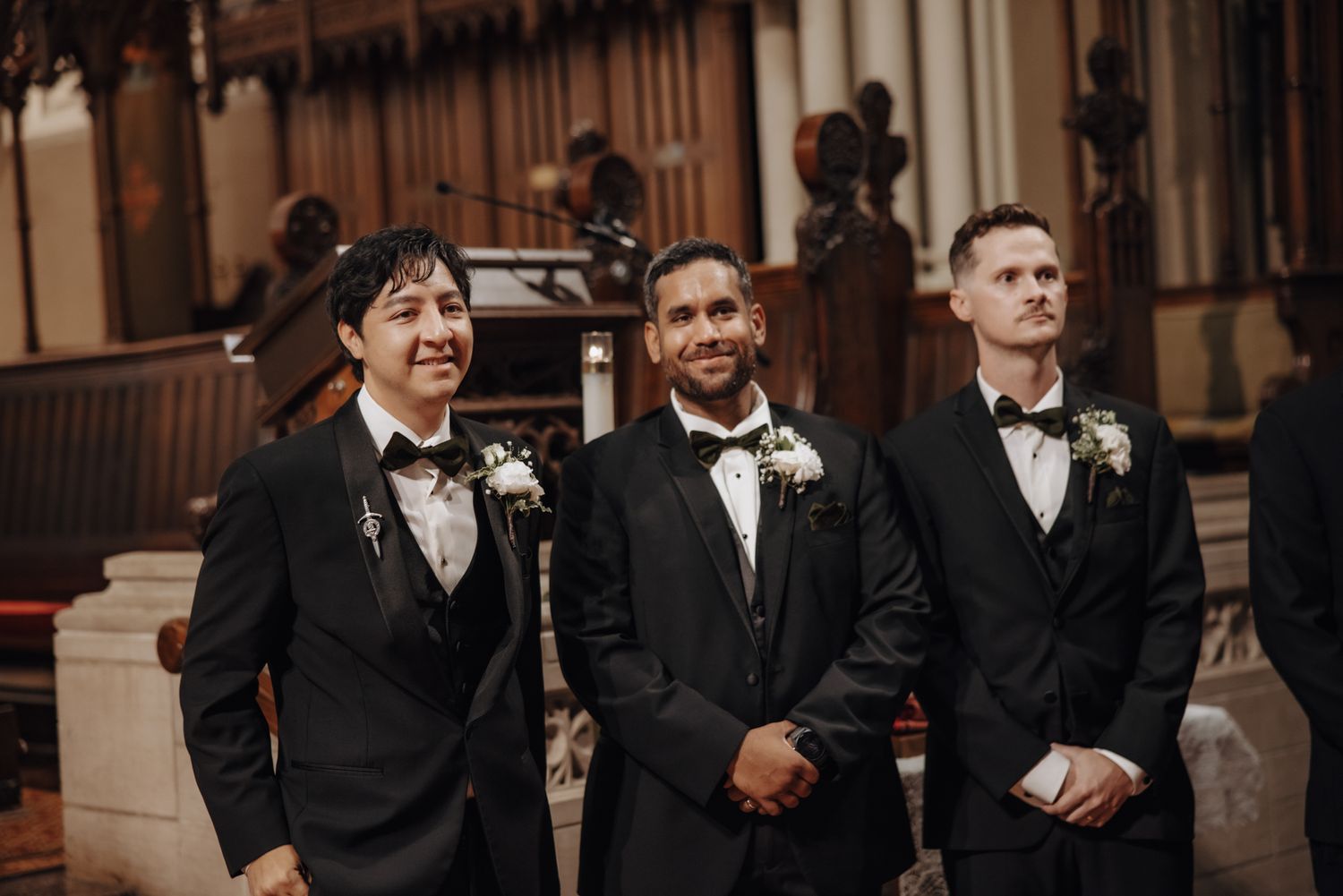 Three men in black tuxedos with bow ties and white boutonnieres stand smiling at the front of a church during a wedding ceremony.