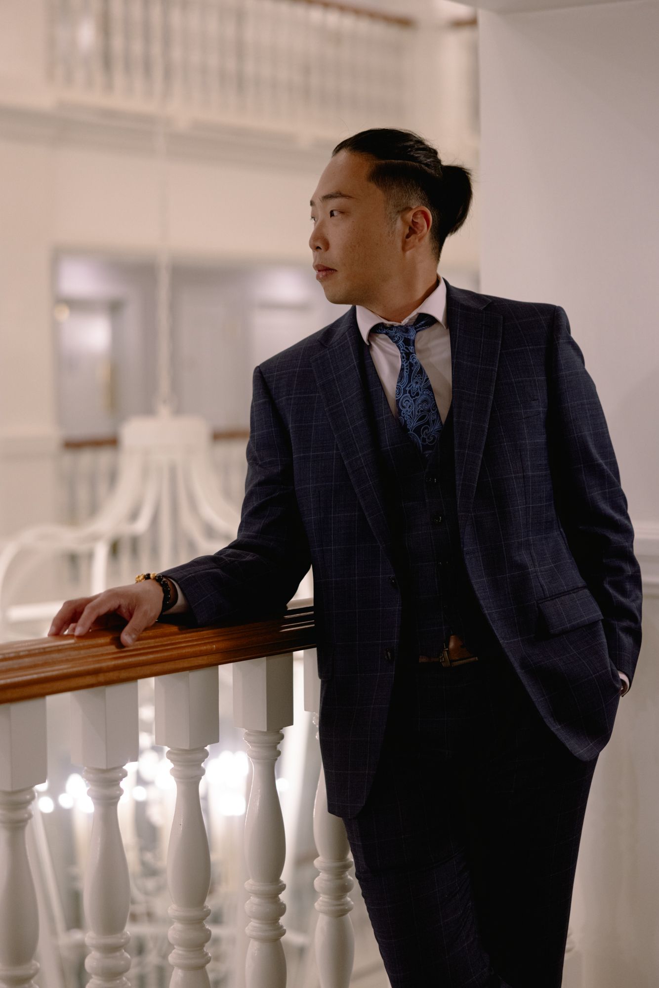 Asian groom in navy plaid three-piece suit with blue tie leaning on white balustrade railing.