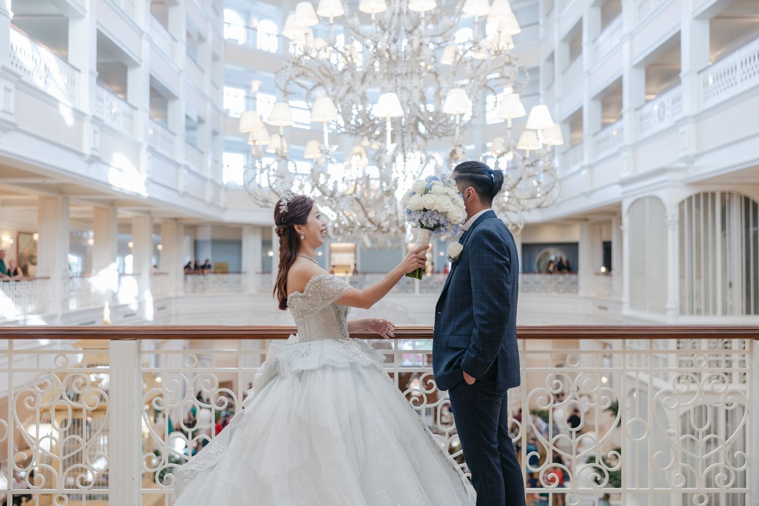 Bride and groom share a romantic moment on a balcony beneath a grand crystal chandelier in an elegant white venue.