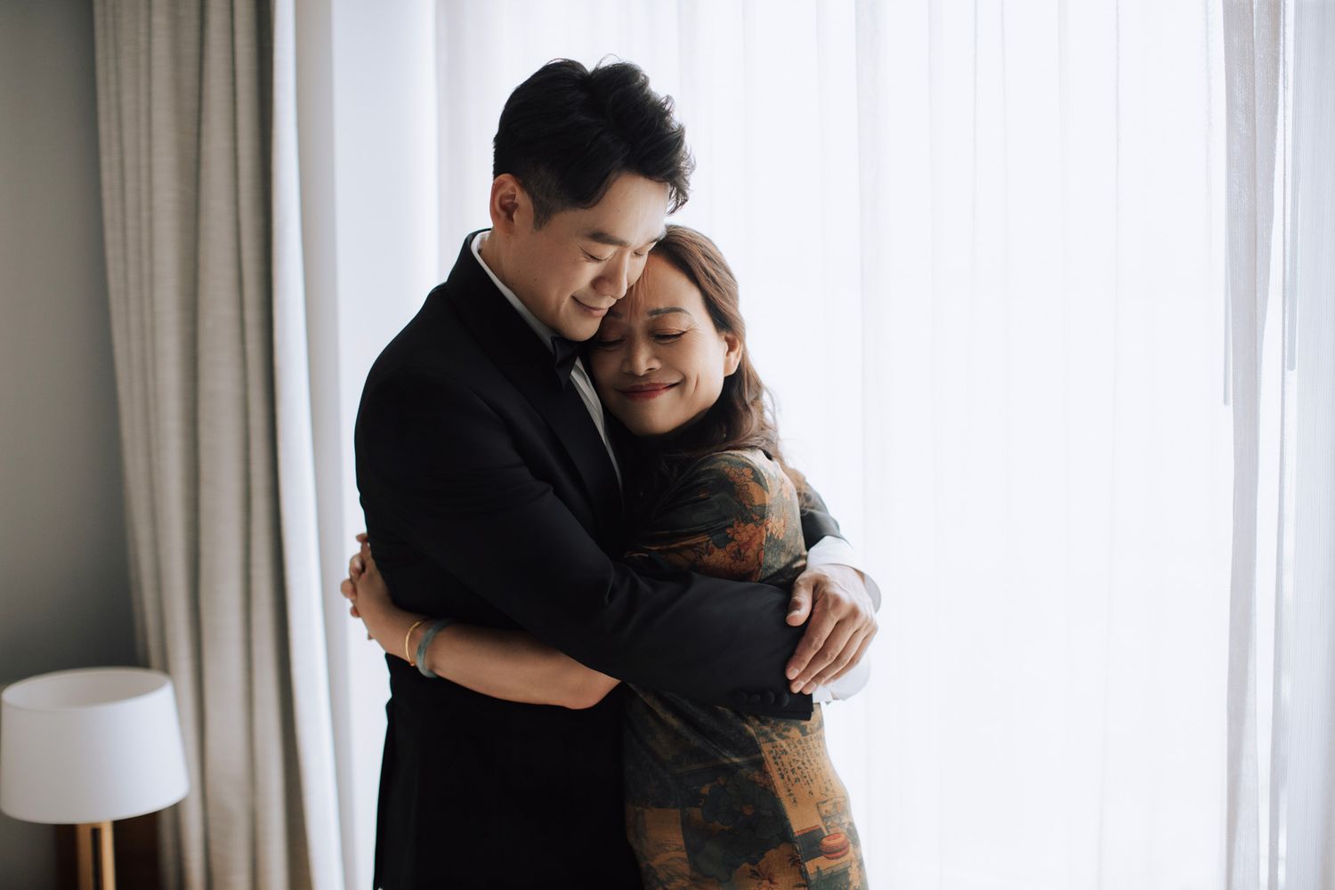 Groom in black tuxedo sharing tender embrace with mother in floral dress by window.