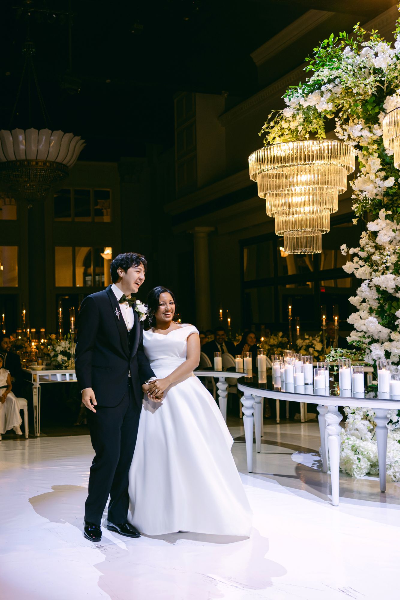 Smiling newlyweds hold hands on a white dance floor beside a floral arrangement with crystal chandeliers and candles.