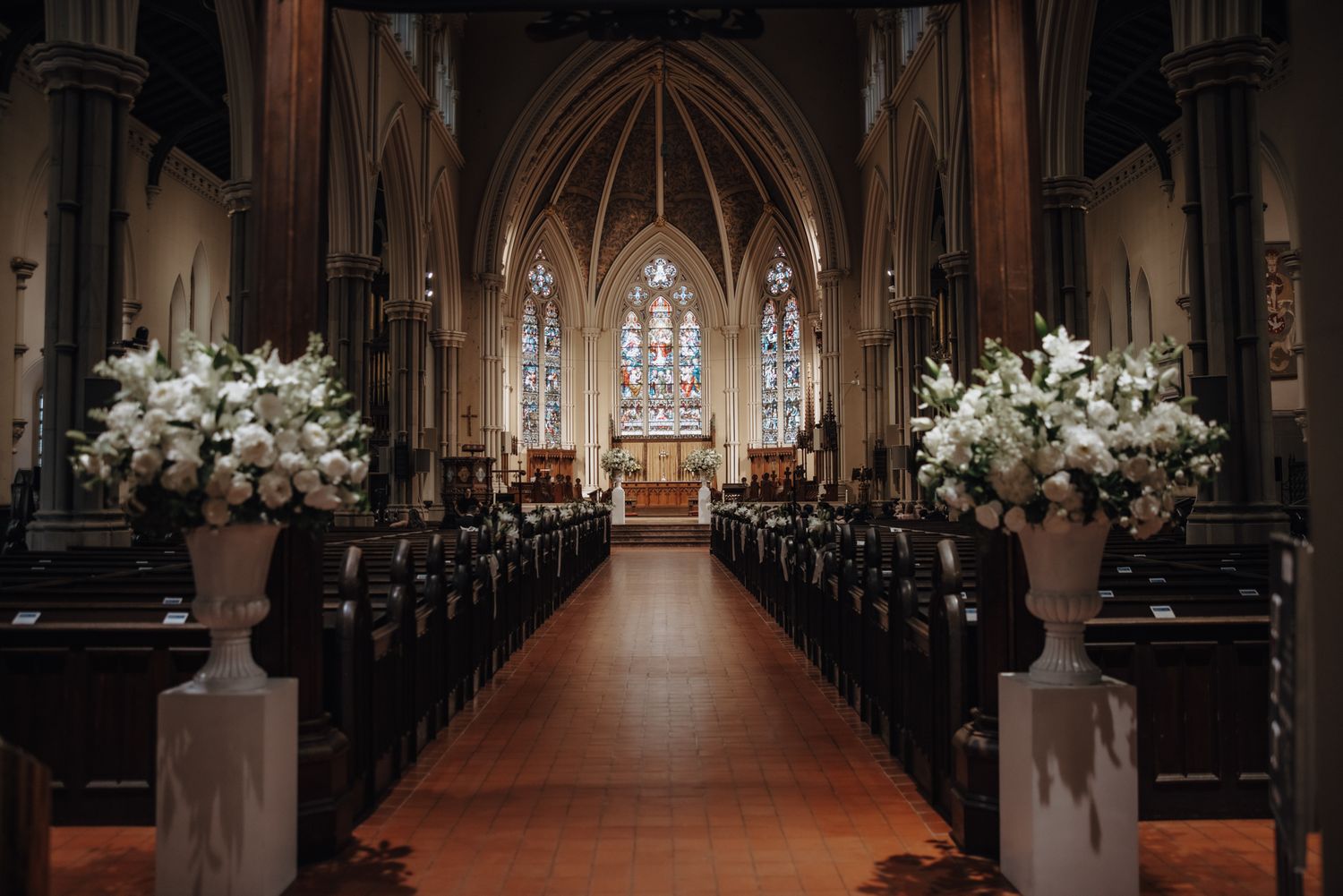 Grand church aisle lined with wooden pews and large white floral arrangements leading to a stained glass window before a wedding.