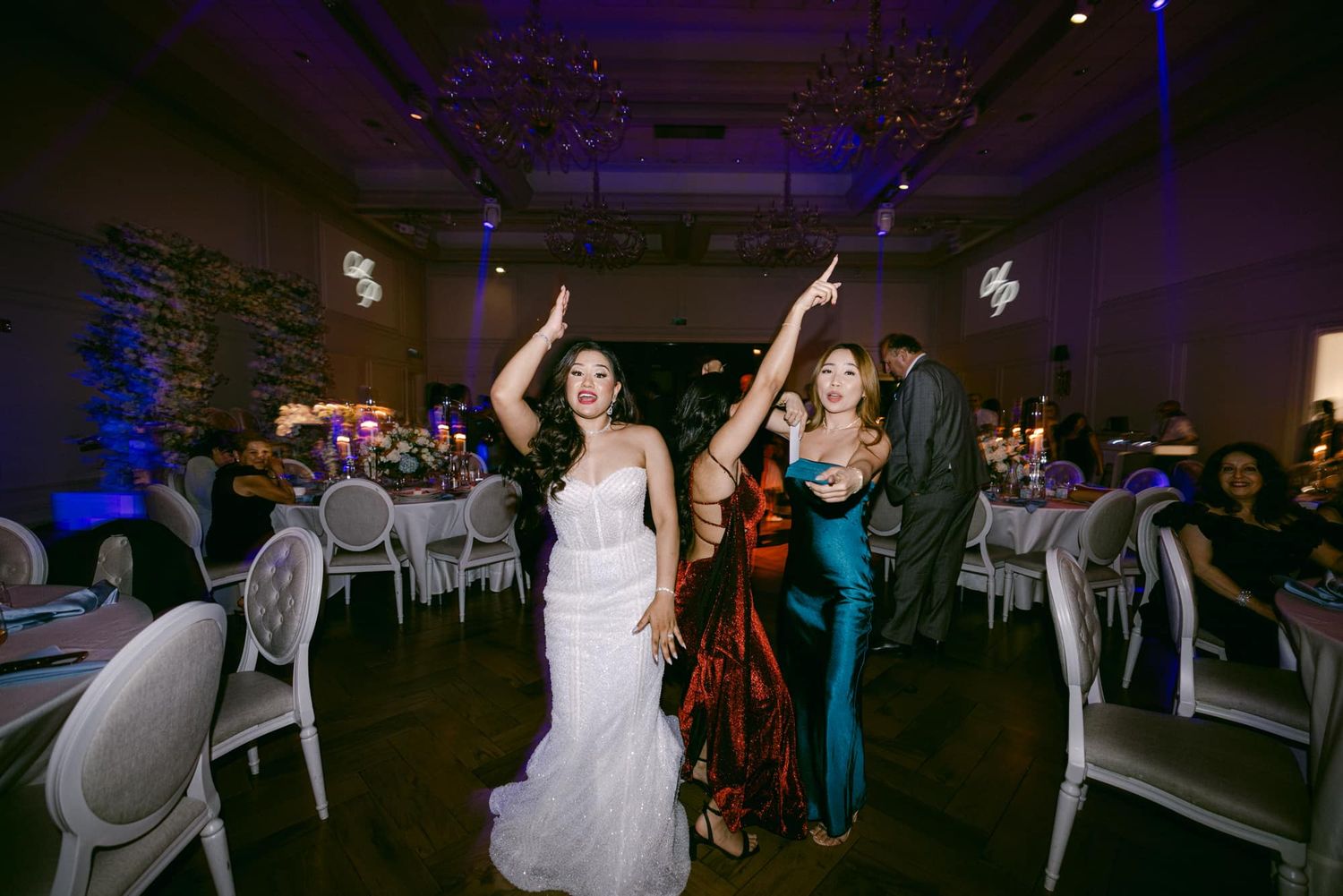 Bride in white gown celebrating with guests raising hands under purple lighting at wedding reception.