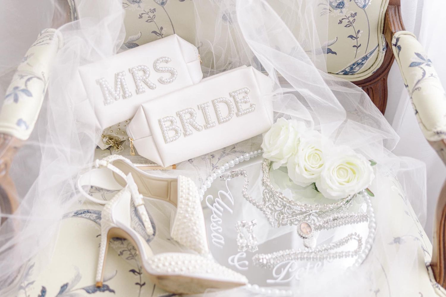 Bridal flat lay featuring white heels, MRS and BRIDE clutches, veil, and white roses on floral chair.