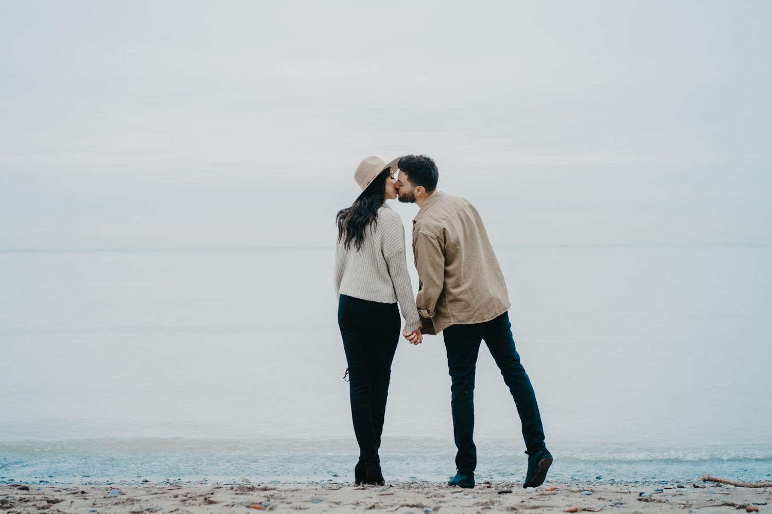 Couple sharing a romantic kiss on a serene beach, holding hands with calm ocean waters in the background.