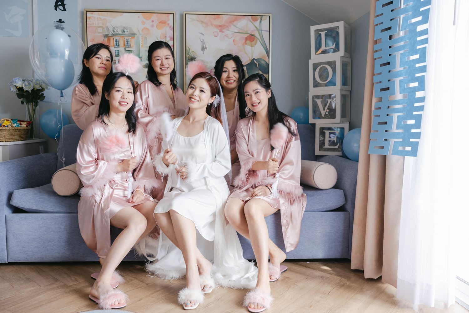 A bride in white robe poses with five bridesmaids in pink robes on a couch, with a LOVE sign in the background.