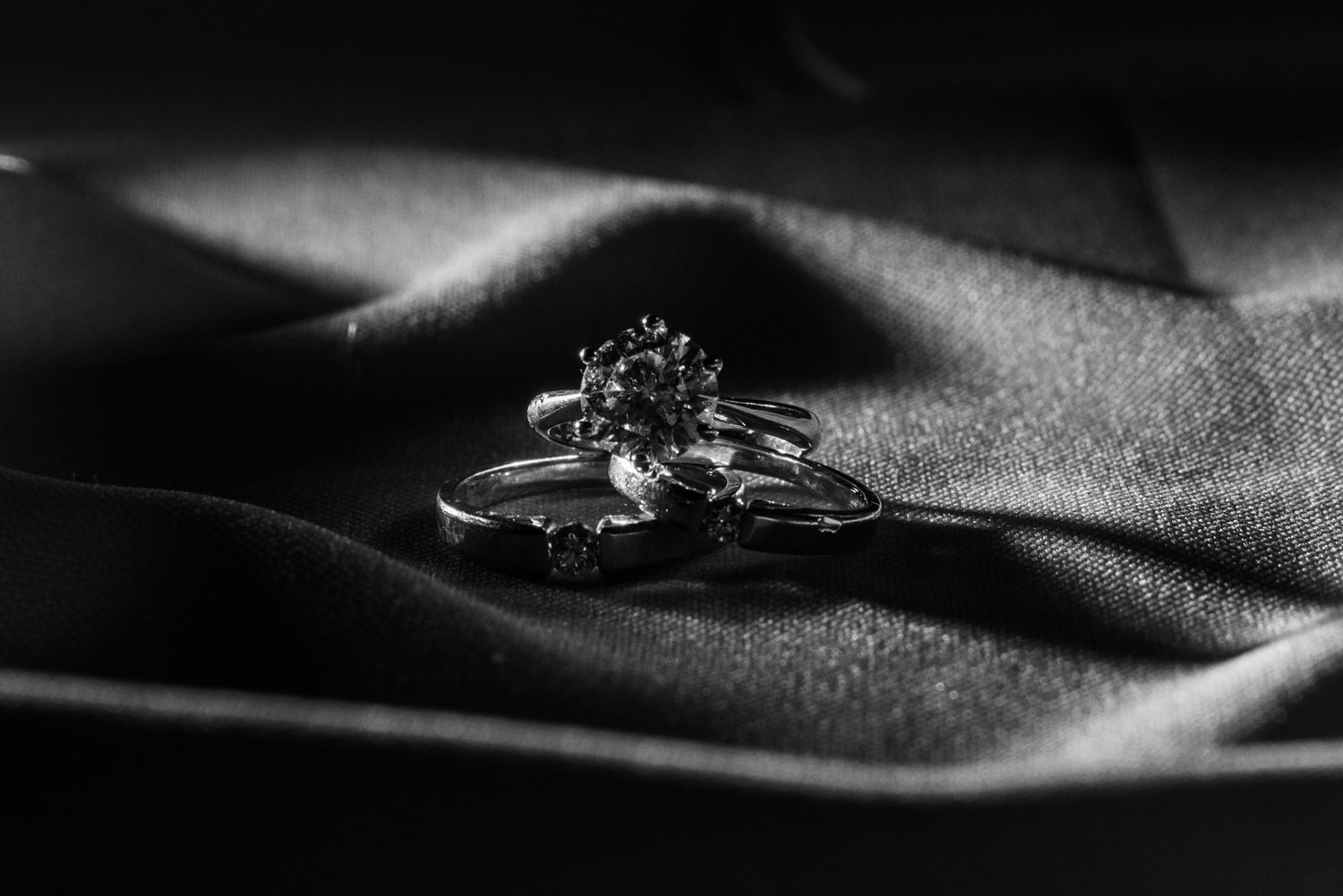Black and white close-up of elegant diamond wedding and engagement rings on silk fabric.