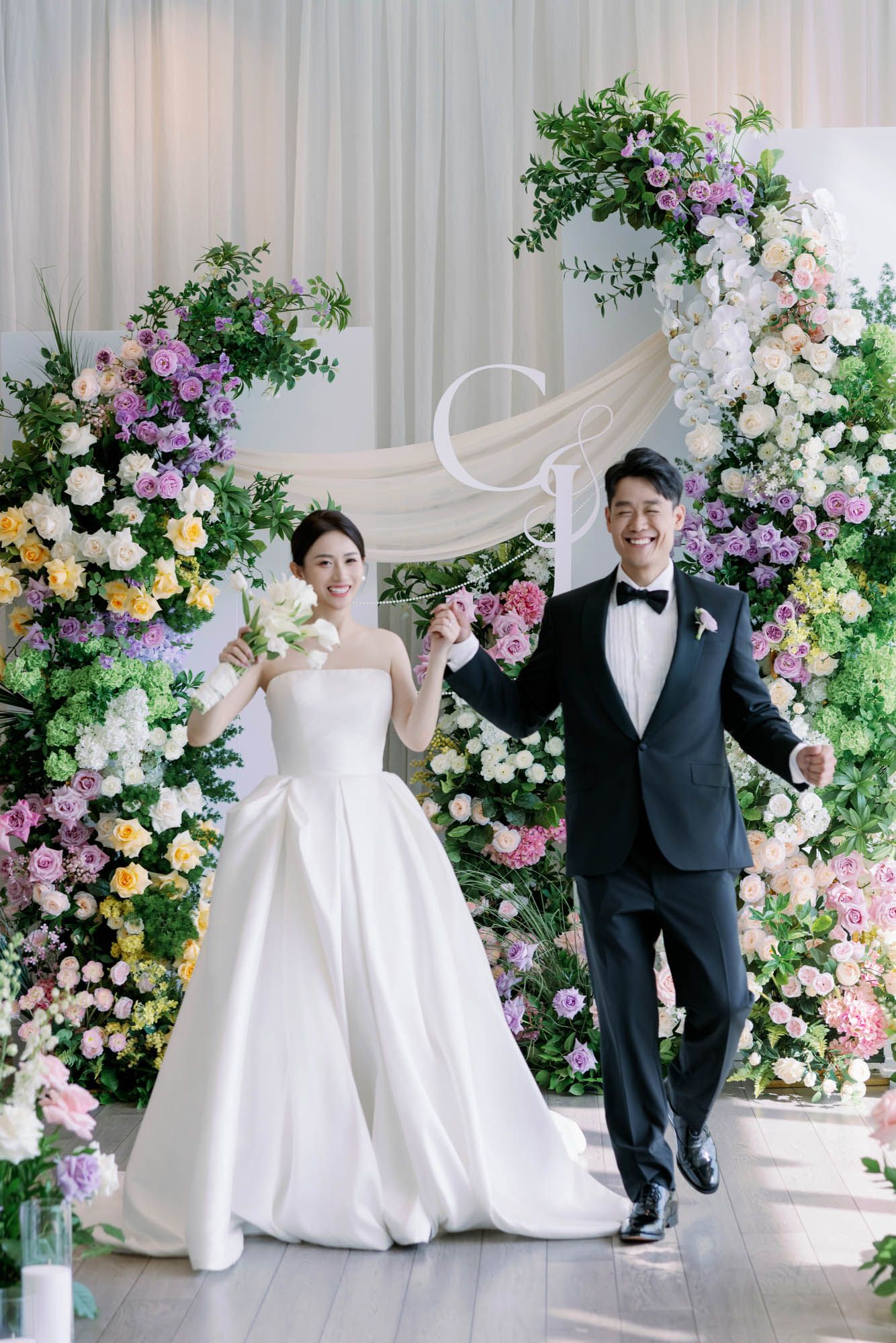 Smiling bride and groom holding hands walking before an elegant flower-covered wedding arch backdrop.