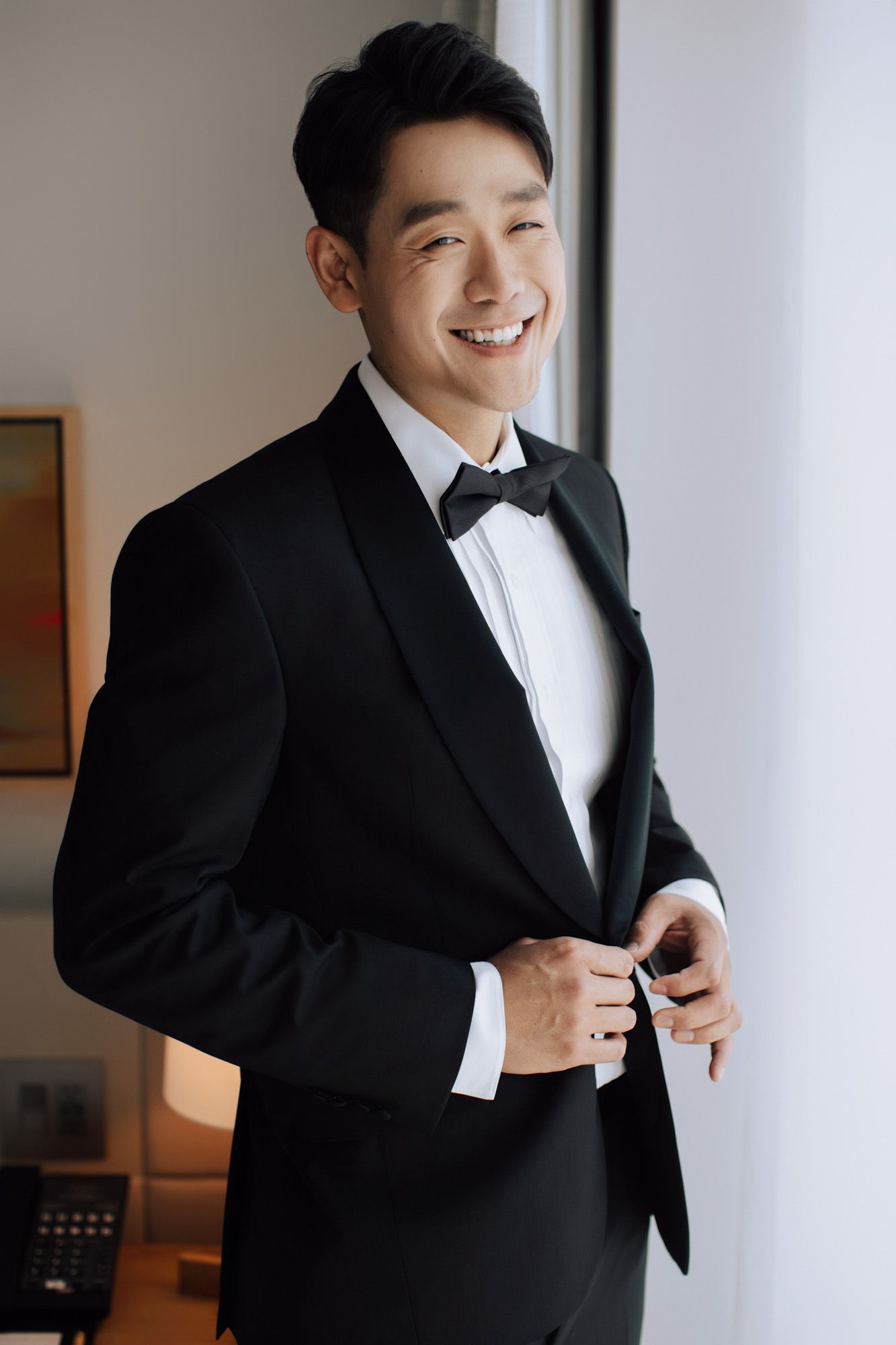 Happy Asian groom in black tuxedo buttoning jacket while smiling near bright window.