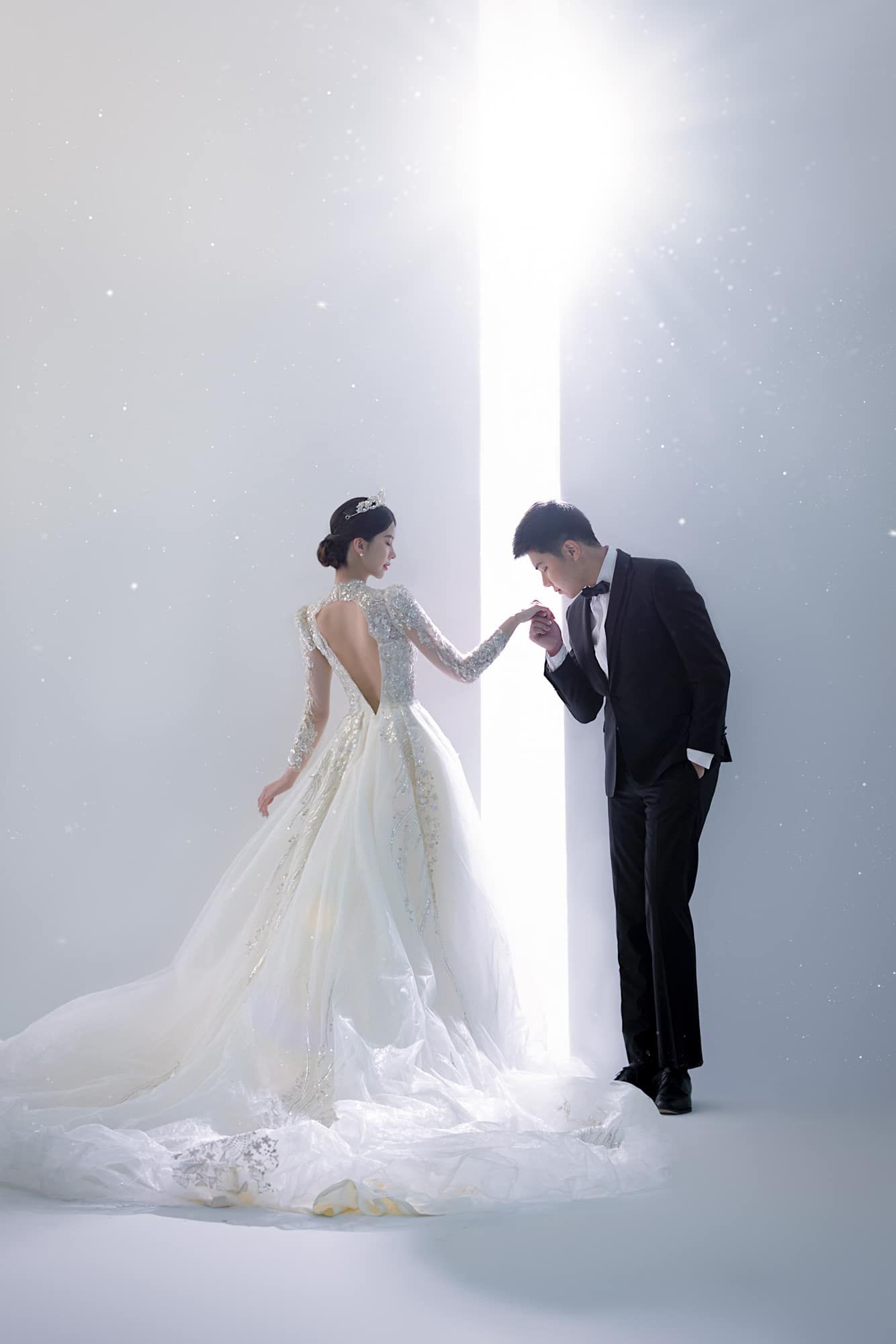 A bride in a white gown and groom in a black tuxedo hold hands before a glowing light in an elegant wedding photo.