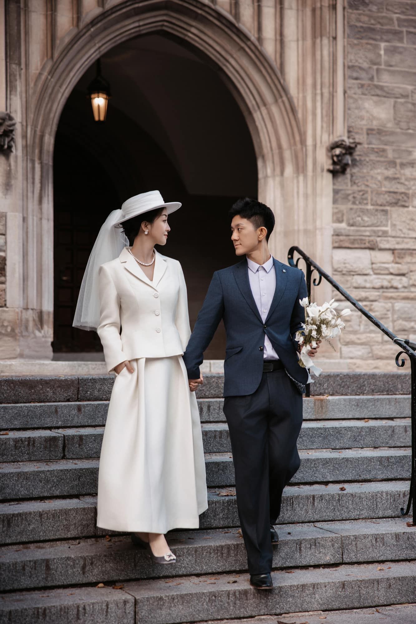 Elegant bride in white coat dress and hat walking down stone church steps with suited groom.