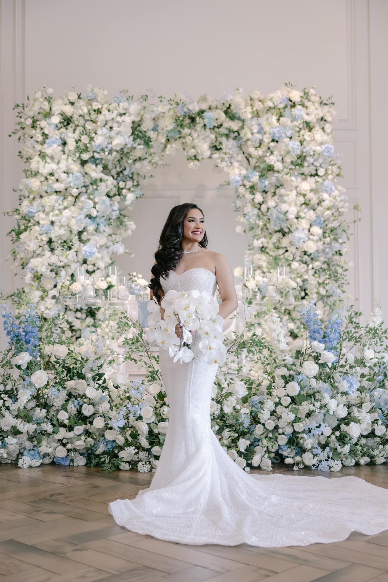 Bride in fitted white gown poses before stunning blue and white floral arch backdrop.