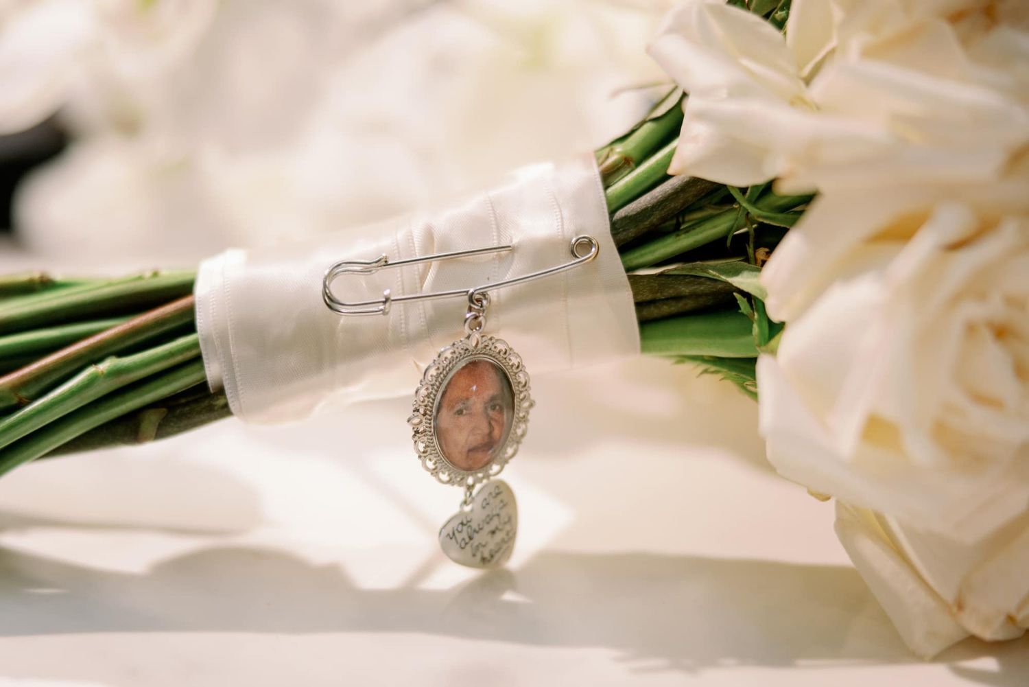 Bridal bouquet charm featuring a vintage photo locket and heart pendant pinned with a safety pin to white roses.