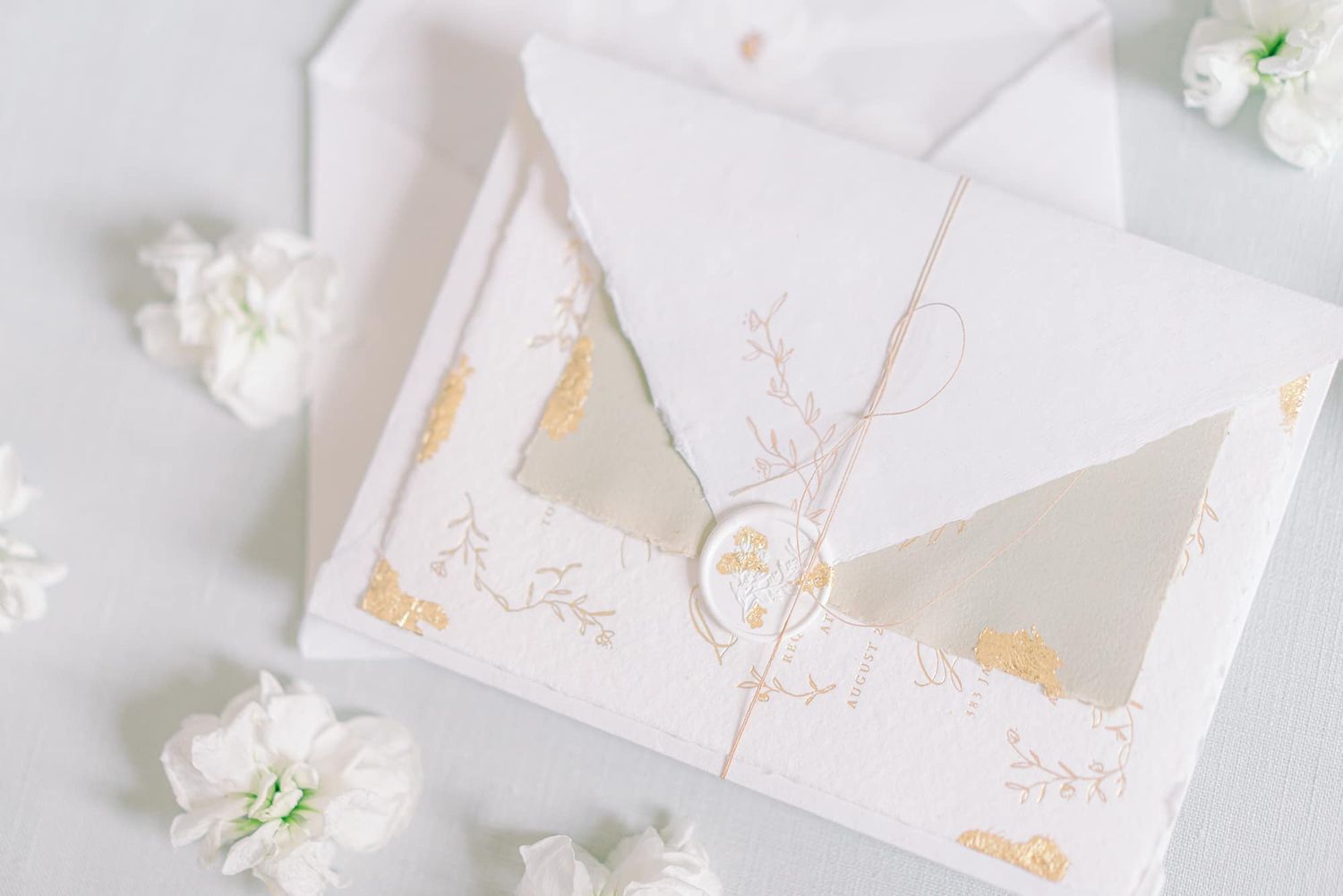 Elegant white wedding invitation with gold foil details and wax seal surrounded by white flowers.