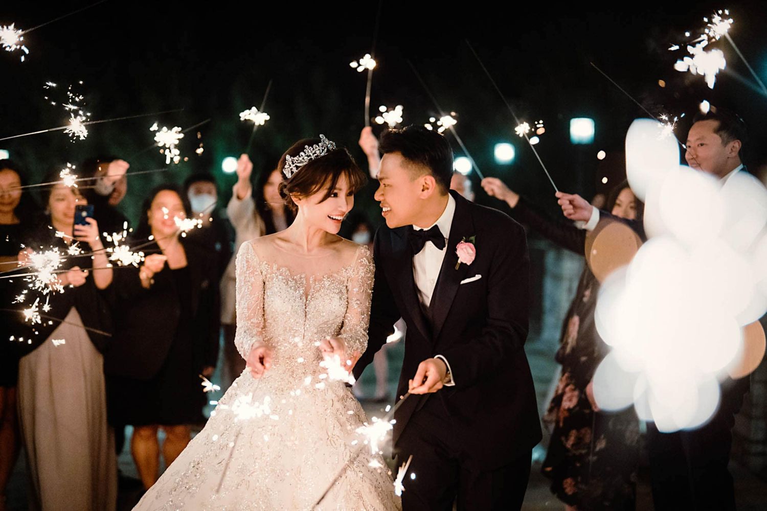 Newlyweds exit celebration surrounded by guests holding sparklers in dazzling nighttime wedding send-off.