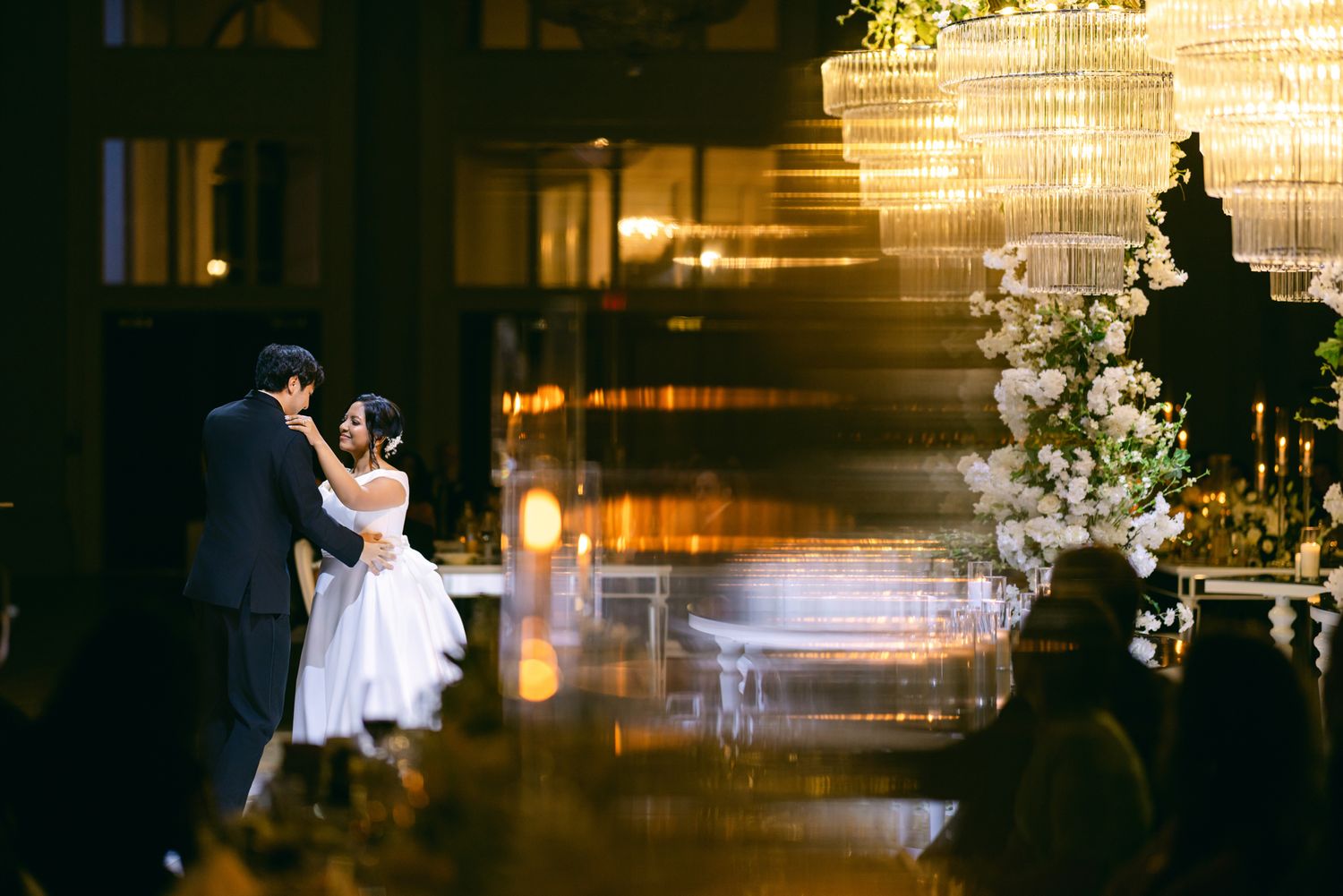 A bride and groom share a romantic dance in a grand ballroom with crystal chandeliers and white floral arrangements.