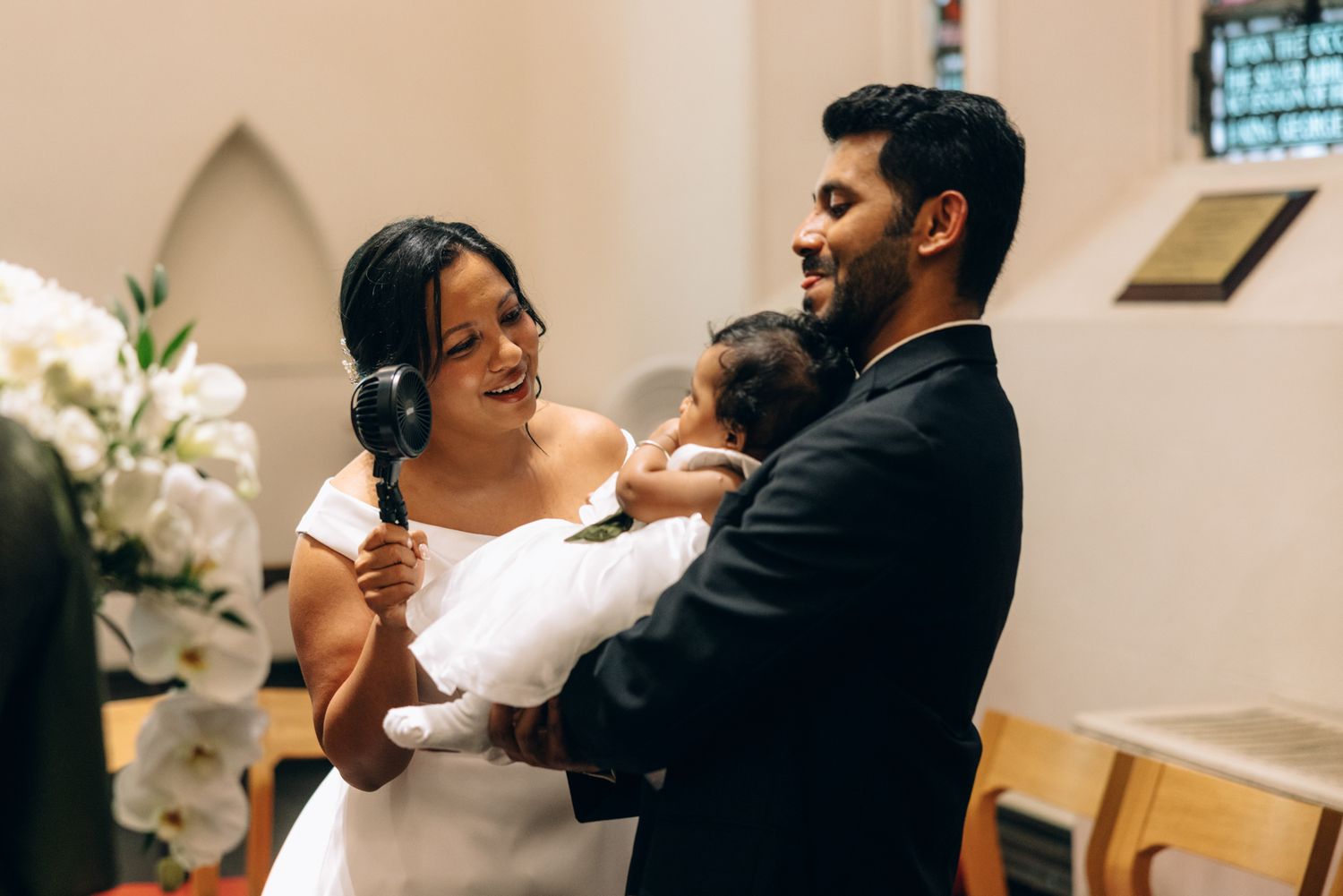 Smiling mother holding a baby in a white gown while a bearded man in a dark suit leans close during a church ceremony.
