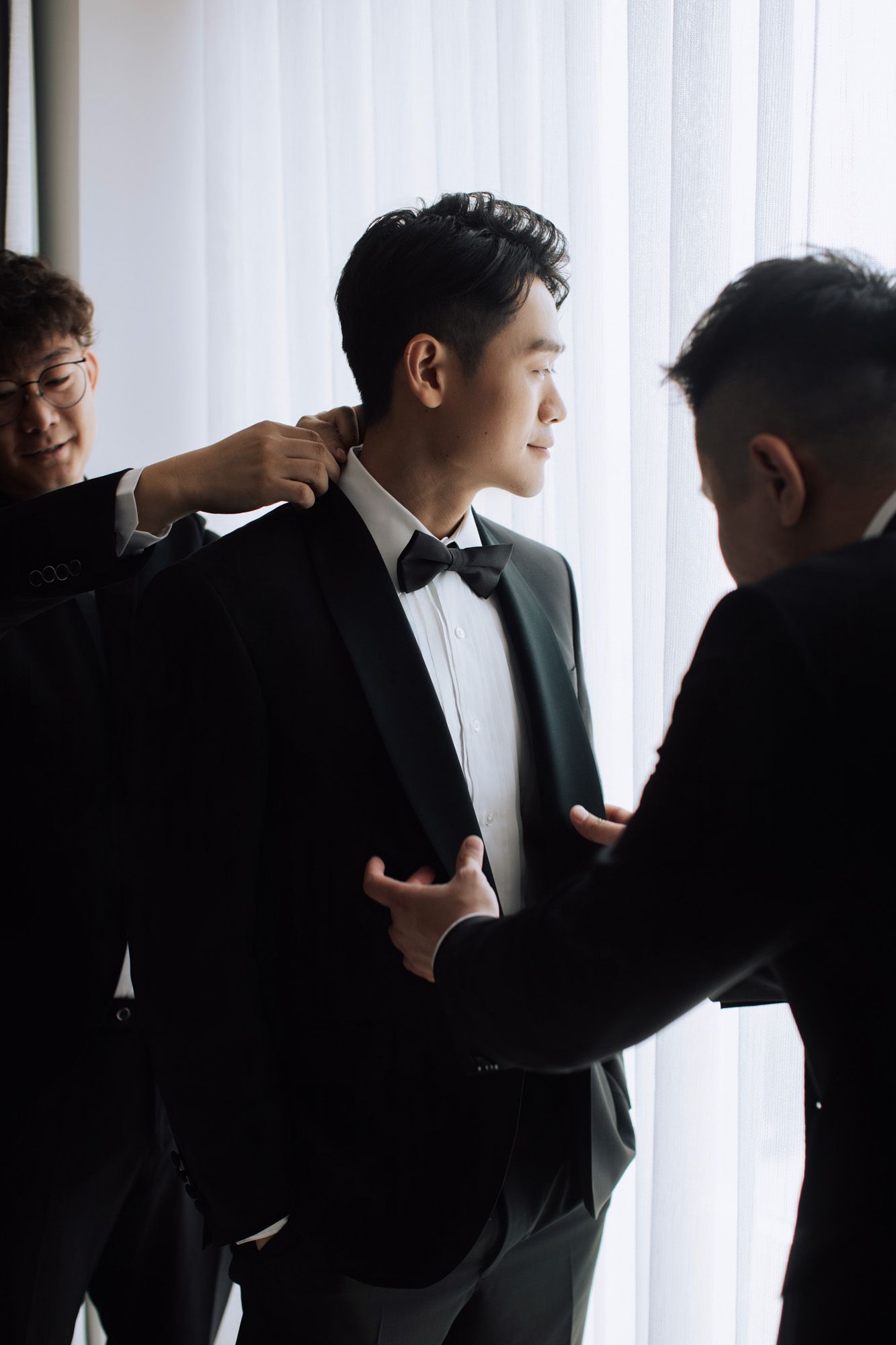 Smiling groom in black tuxedo being helped by two groomsmen near bright curtained window.