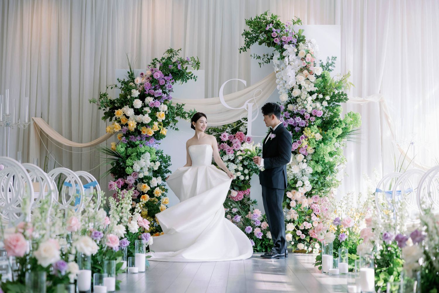 Bride in elegant ballgown stands with groom beneath a colorful floral arch at indoor wedding.