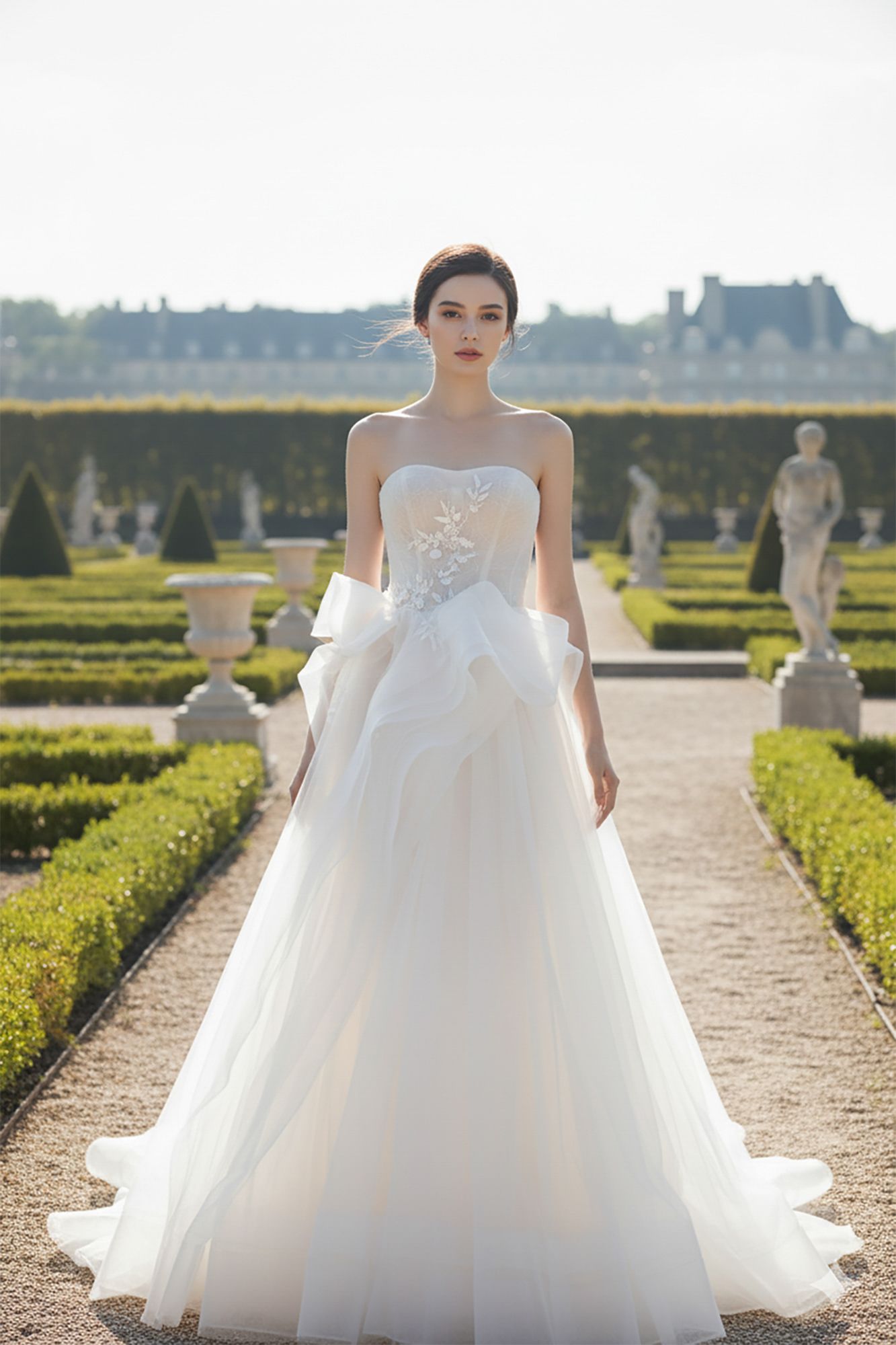 Bride in strapless white ballgown with lace bodice and tulle skirt posing in formal garden with hedges and statues.