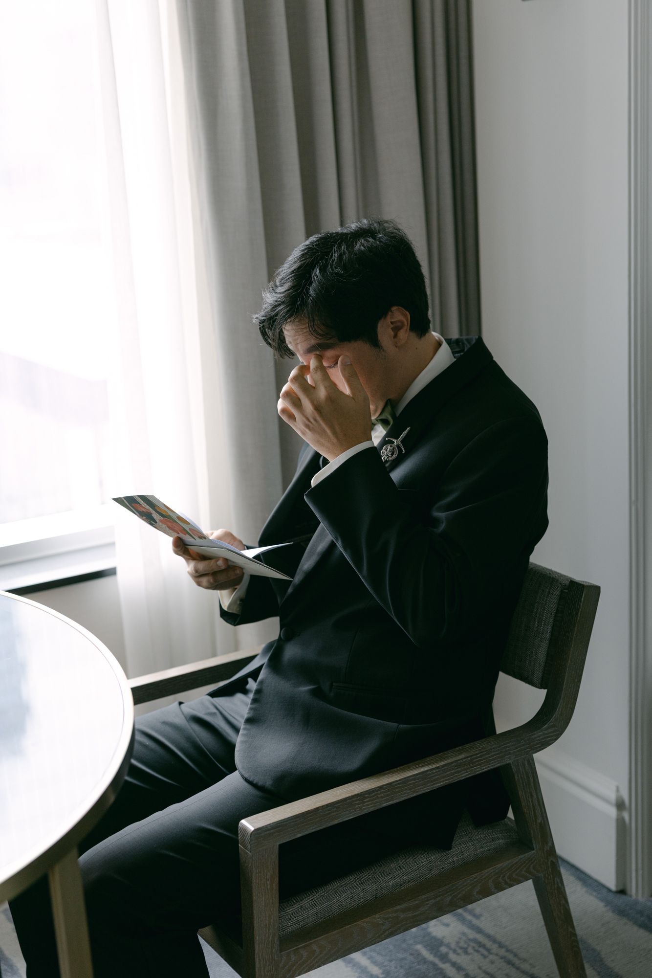 Emotional groom in a black suit sits in a wooden chair by a sunlit window, wiping his eyes while reading a card on his wedding day.