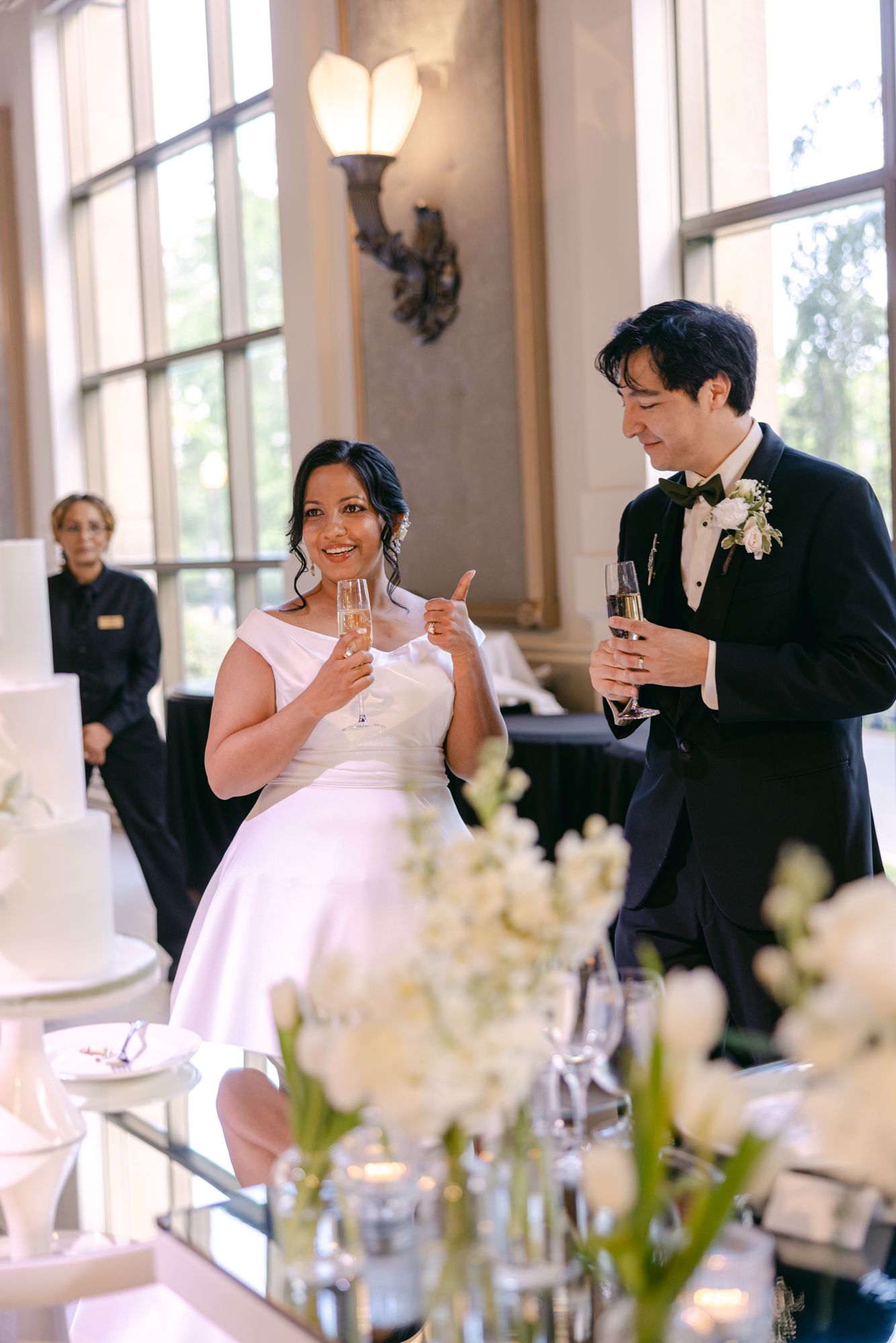 Smiling bride in a white gown and groom in a black tuxedo toast with champagne beside white flowers and a wedding cake at their reception.