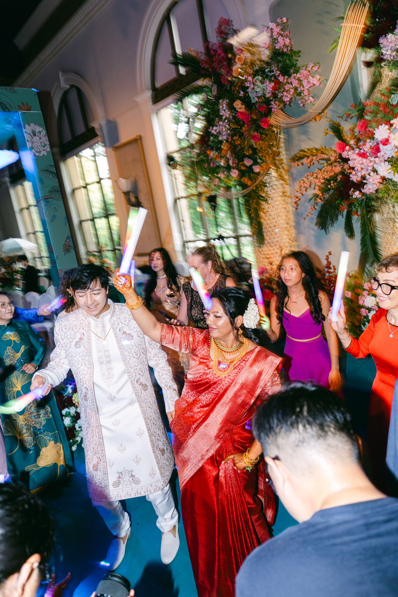A groom in a white sherwani and a bride in a red sari dance with glow sticks as guests cheer at a floral decorated wedding reception.