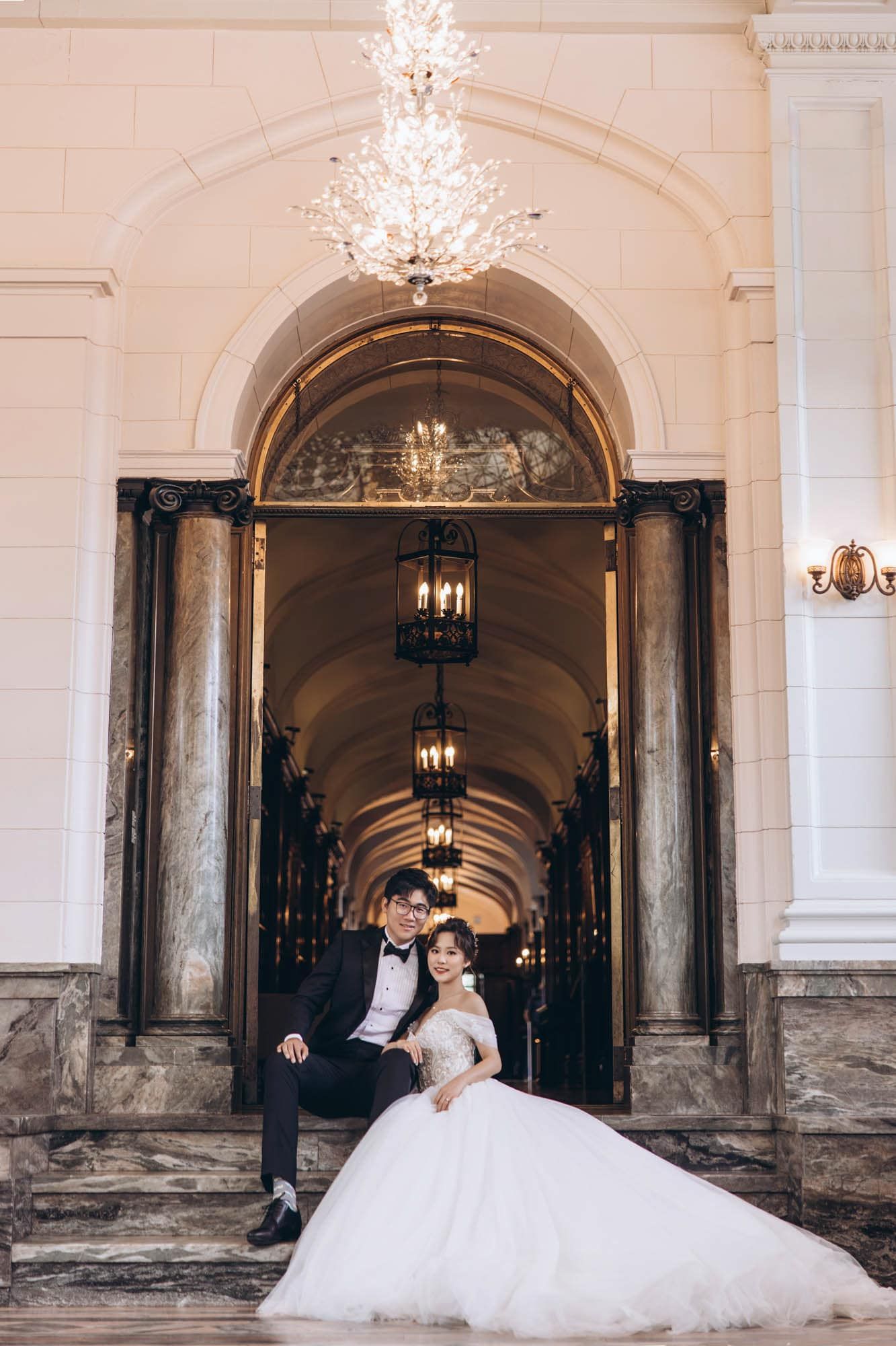 Bride and groom pose elegantly in a marble archway corridor with crystal chandeliers.