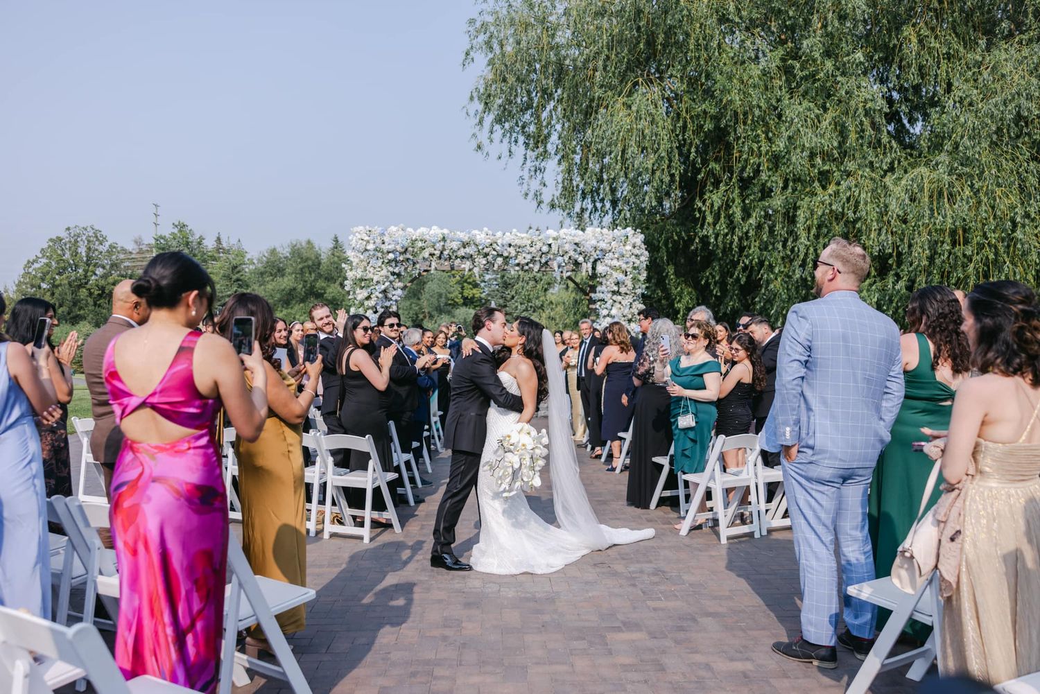 Newlyweds share spontaneous kiss while exiting outdoor ceremony aisle with cheering guests.
