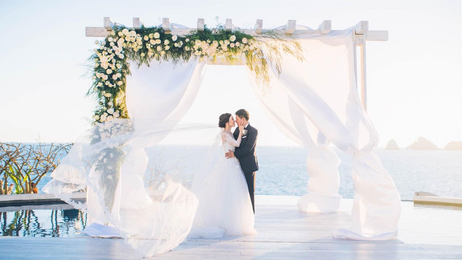 Couple shares first kiss under white floral arch with flowing drapes overlooking ocean at sunset.