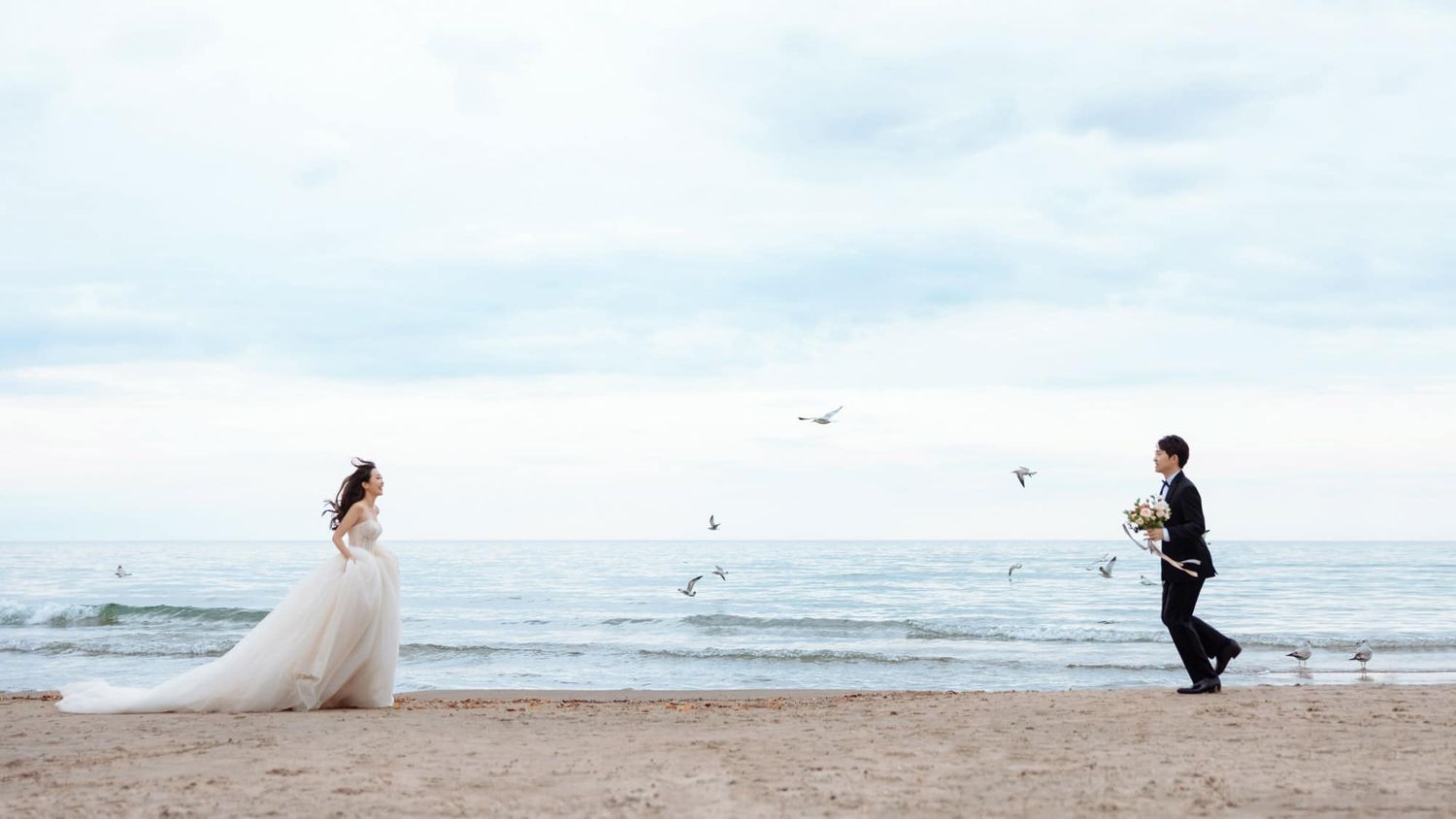 Bride and groom walk toward each other on a serene beach with seagulls flying overhead.