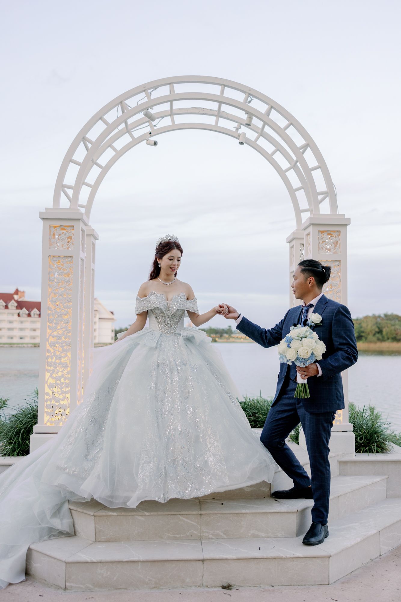 Bride in silver ballgown and groom in navy suit hold hands under white arch by waterfront.