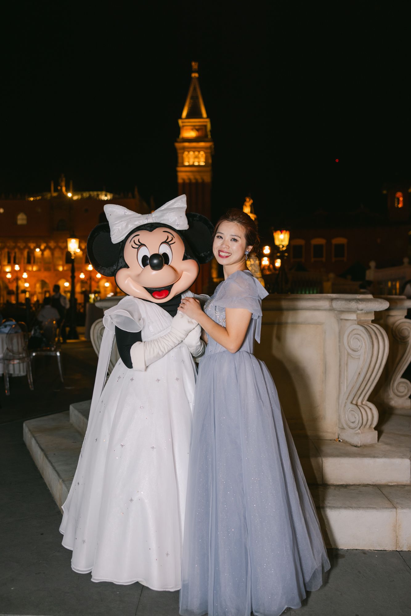 Bride in lavender gown poses with Minnie Mouse in wedding dress against illuminated Disney backdrop.