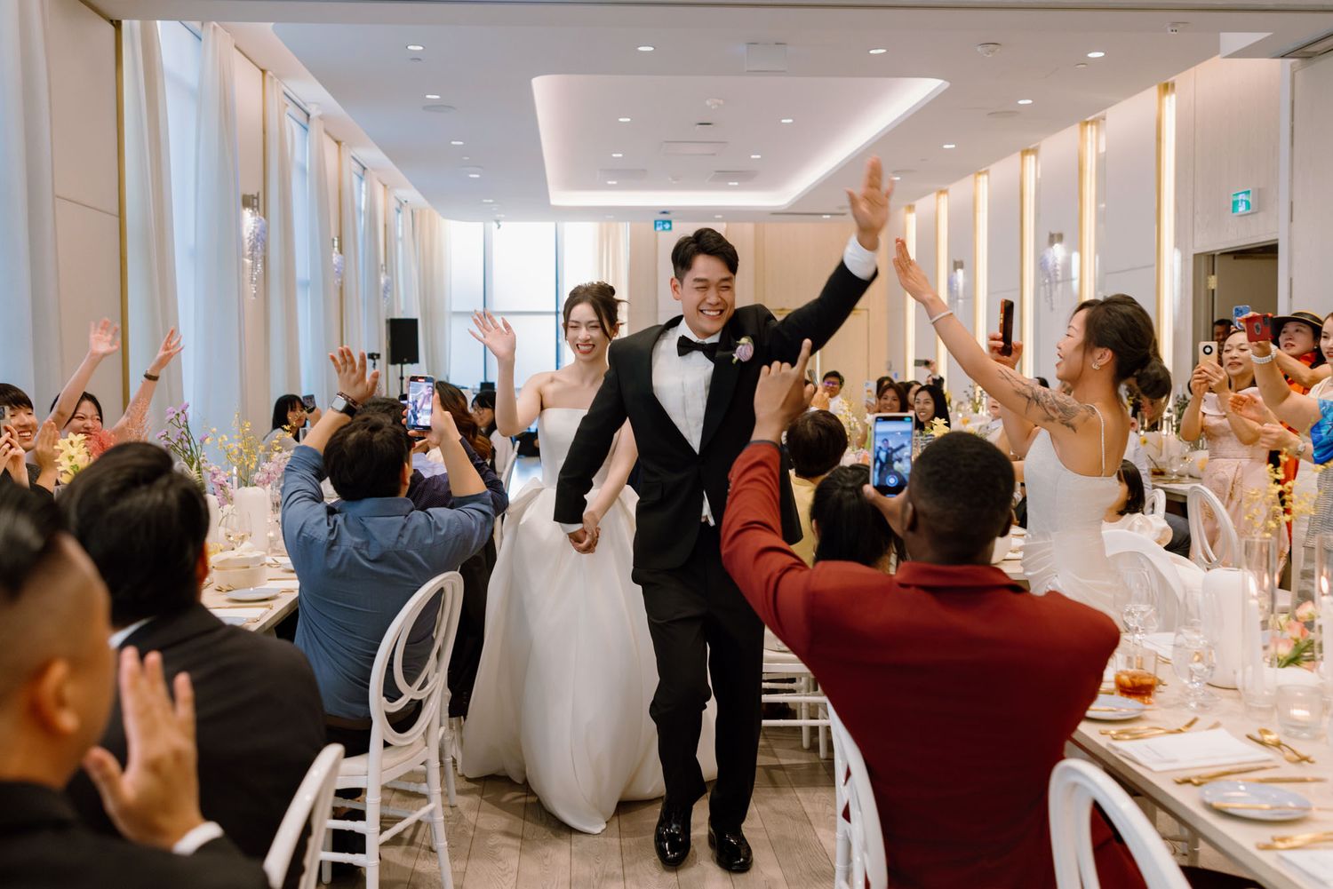Bride and groom joyfully entering wedding reception as guests cheer and raise their hands in celebration.