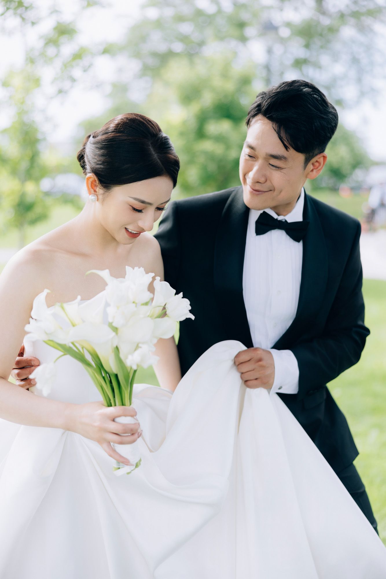 Groom in black tuxedo helps bride with white ball gown and calla lily bouquet outdoors.