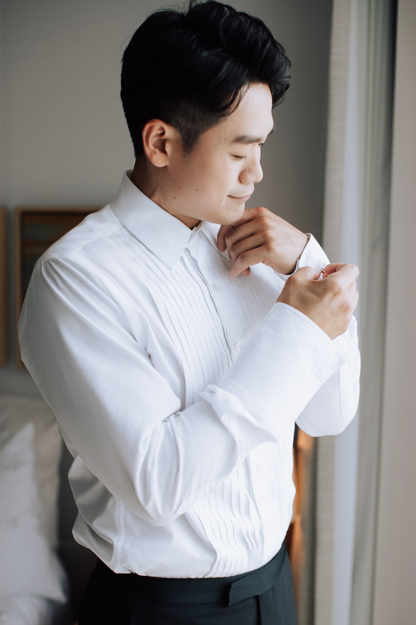Asian groom in white tuxedo shirt fastening cufflinks while getting ready on wedding day.