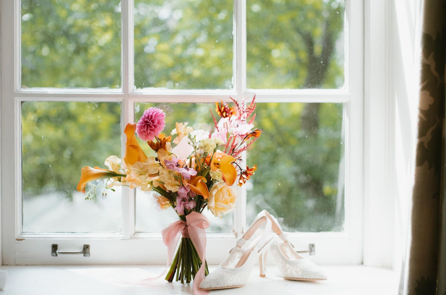Colorful bridal bouquet with pink and orange flowers beside white heels on a windowsill.