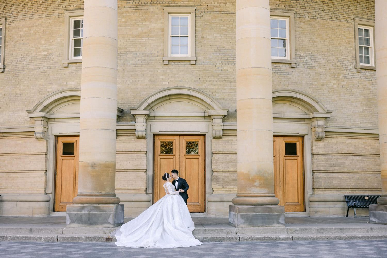 Bride and groom embrace in front of a grand stone church with tall columns and wooden doors on their wedding day.