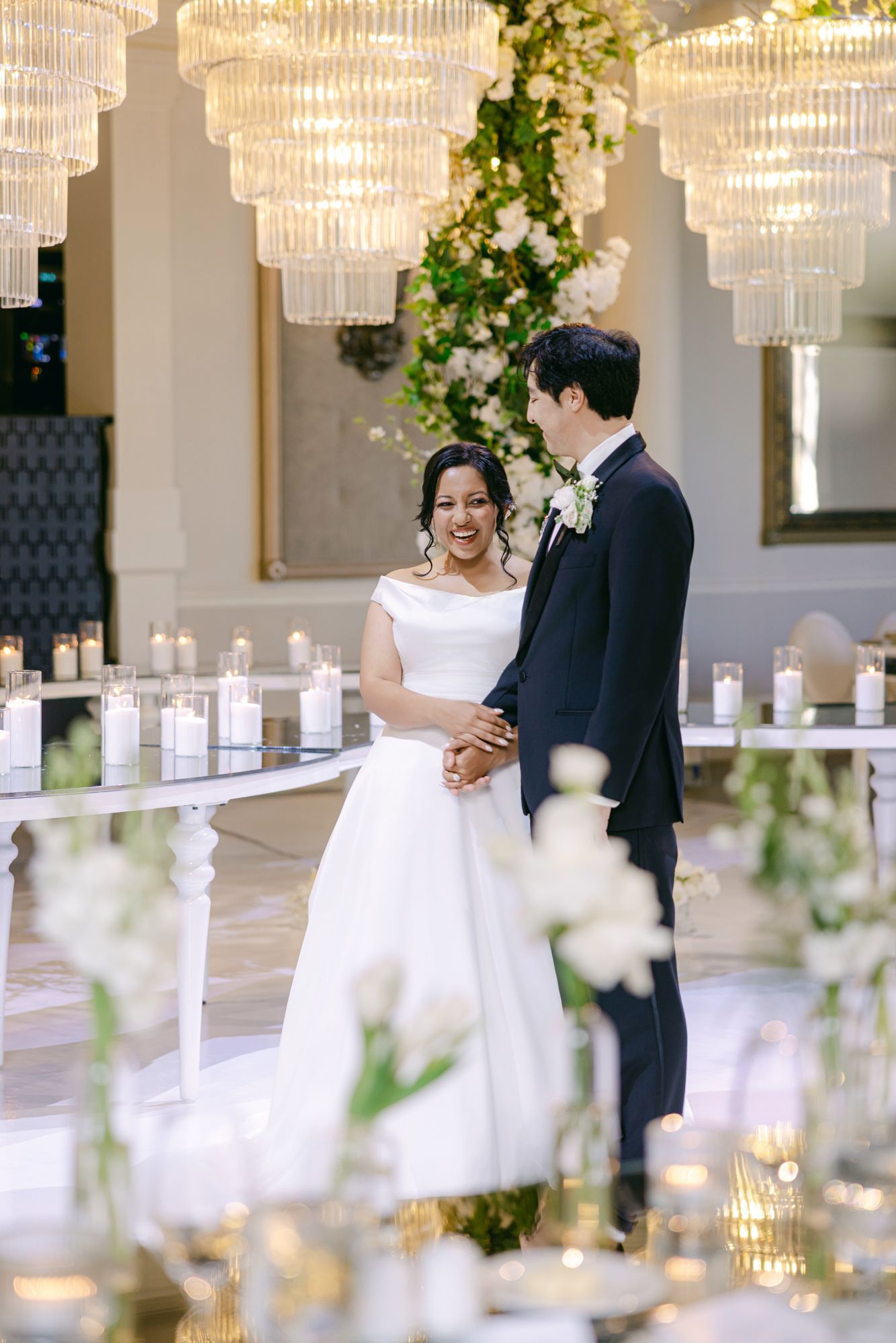 Smiling bride in an off-shoulder white gown holds hands with the groom in a navy suit beside candlelit tables and crystal chandeliers.