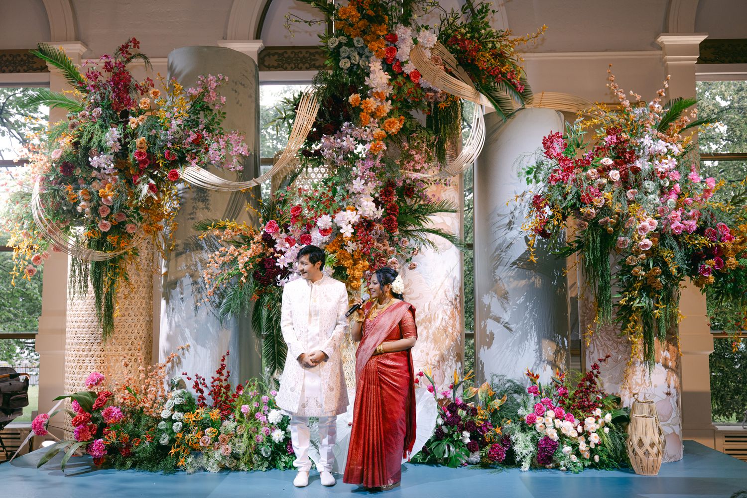 A couple in a white sherwani and a red saree stand together before large pink and orange floral arrangements on a decorated wedding stage.