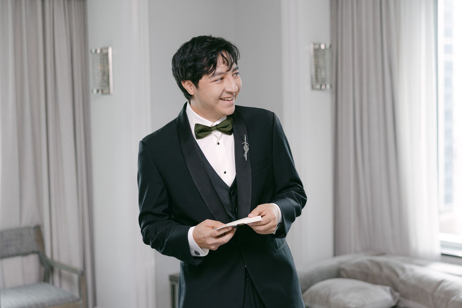 Smiling groom in a black tuxedo and green bow tie glances aside while holding a folded note in a softly lit room with sheer curtains.
