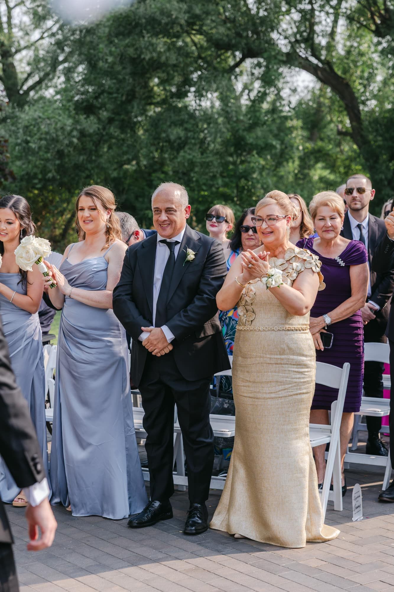 Proud parents and bridesmaids in silver gowns watch wedding ceremony outdoors.