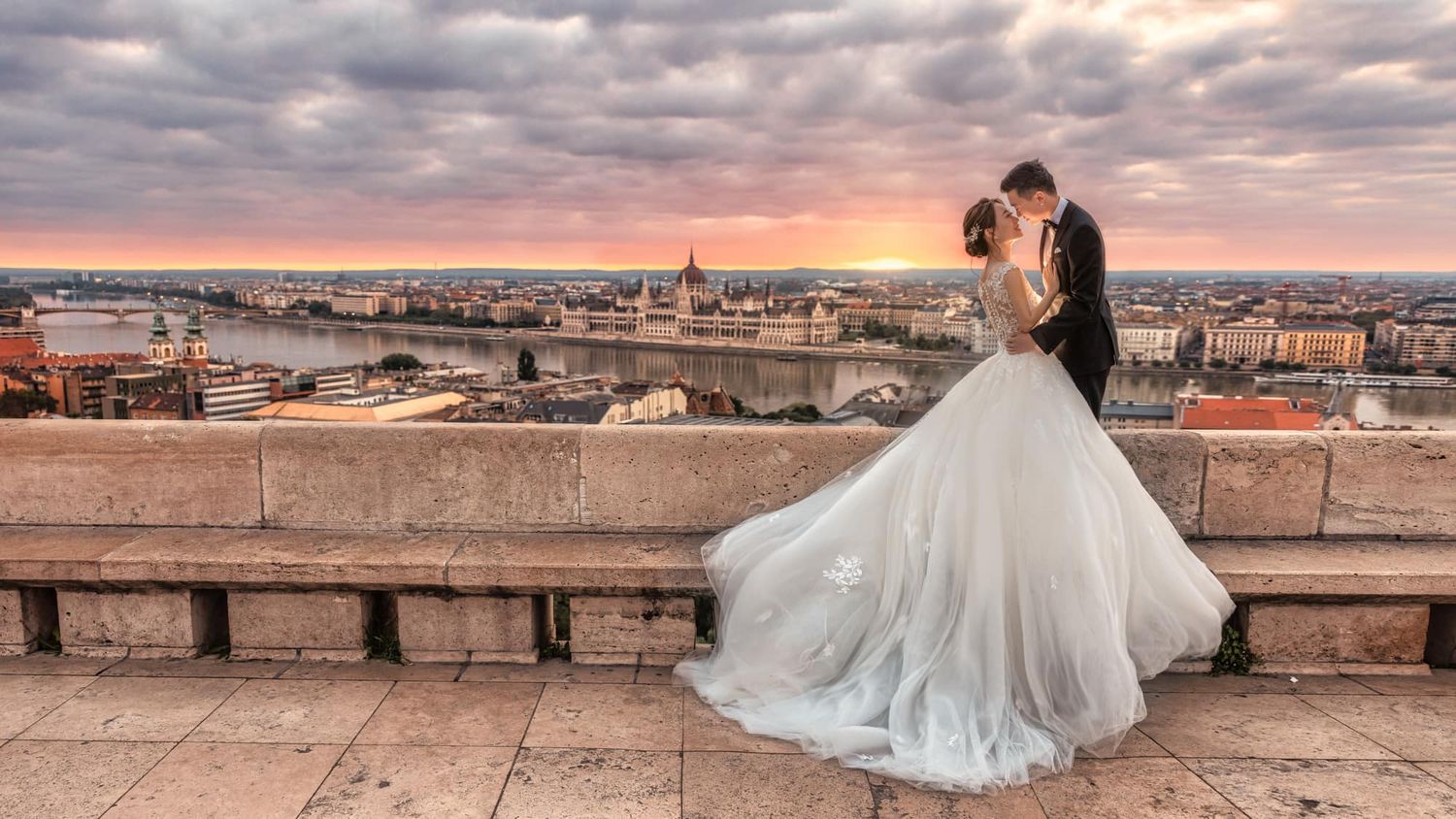 Bride and groom share romantic embrace on scenic overlook at sunset with city skyline and river in background.