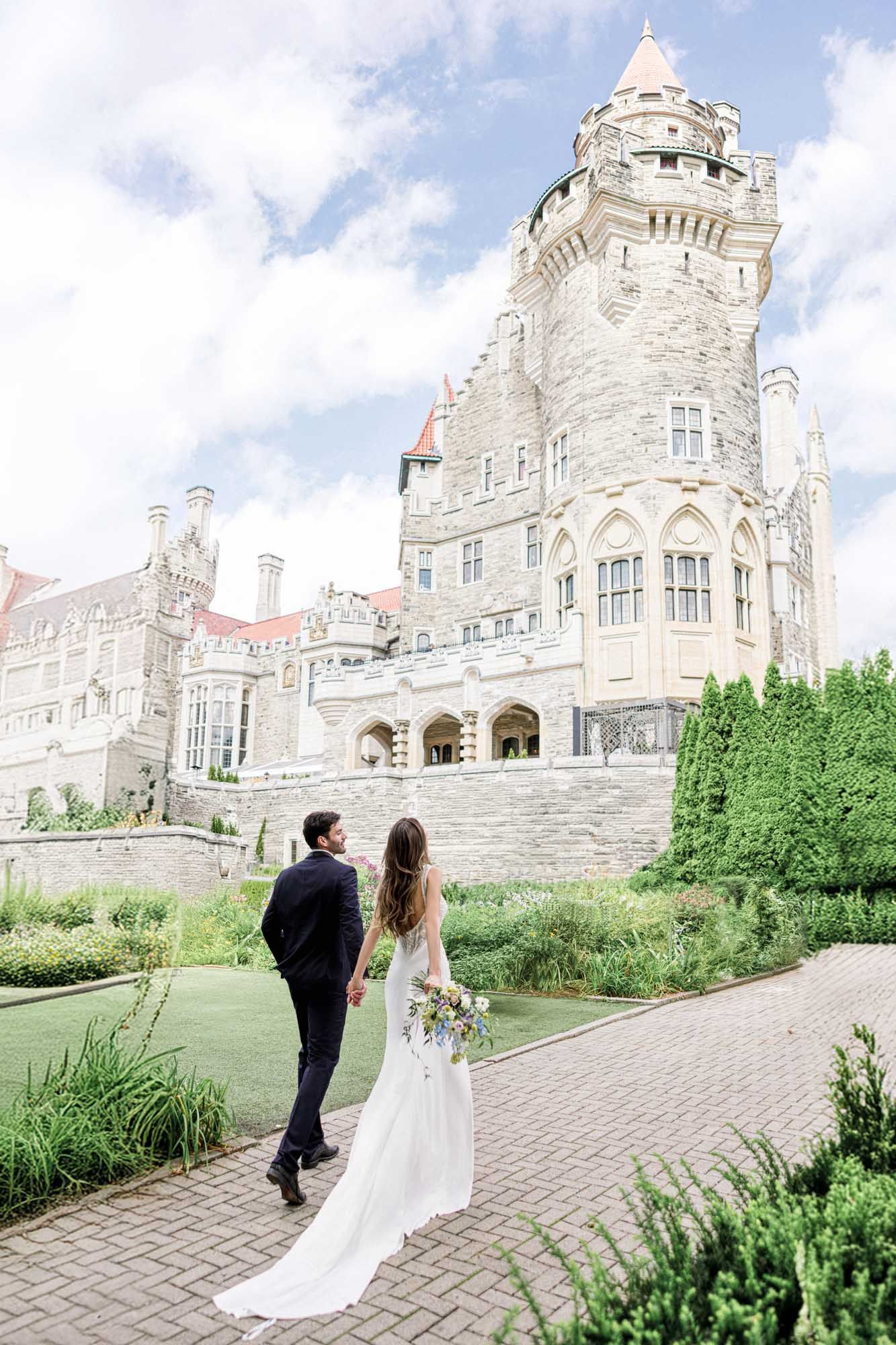 Bride and groom walking hand-in-hand toward a grand stone castle surrounded by lush green gardens.