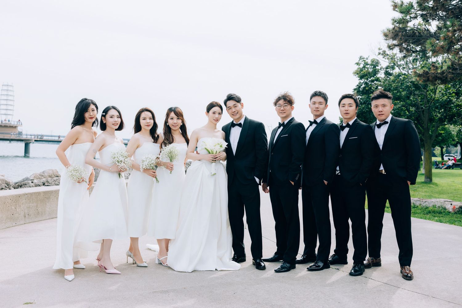 Wedding party of bridesmaids in white and groomsmen in black tuxedos pose by waterfront.
