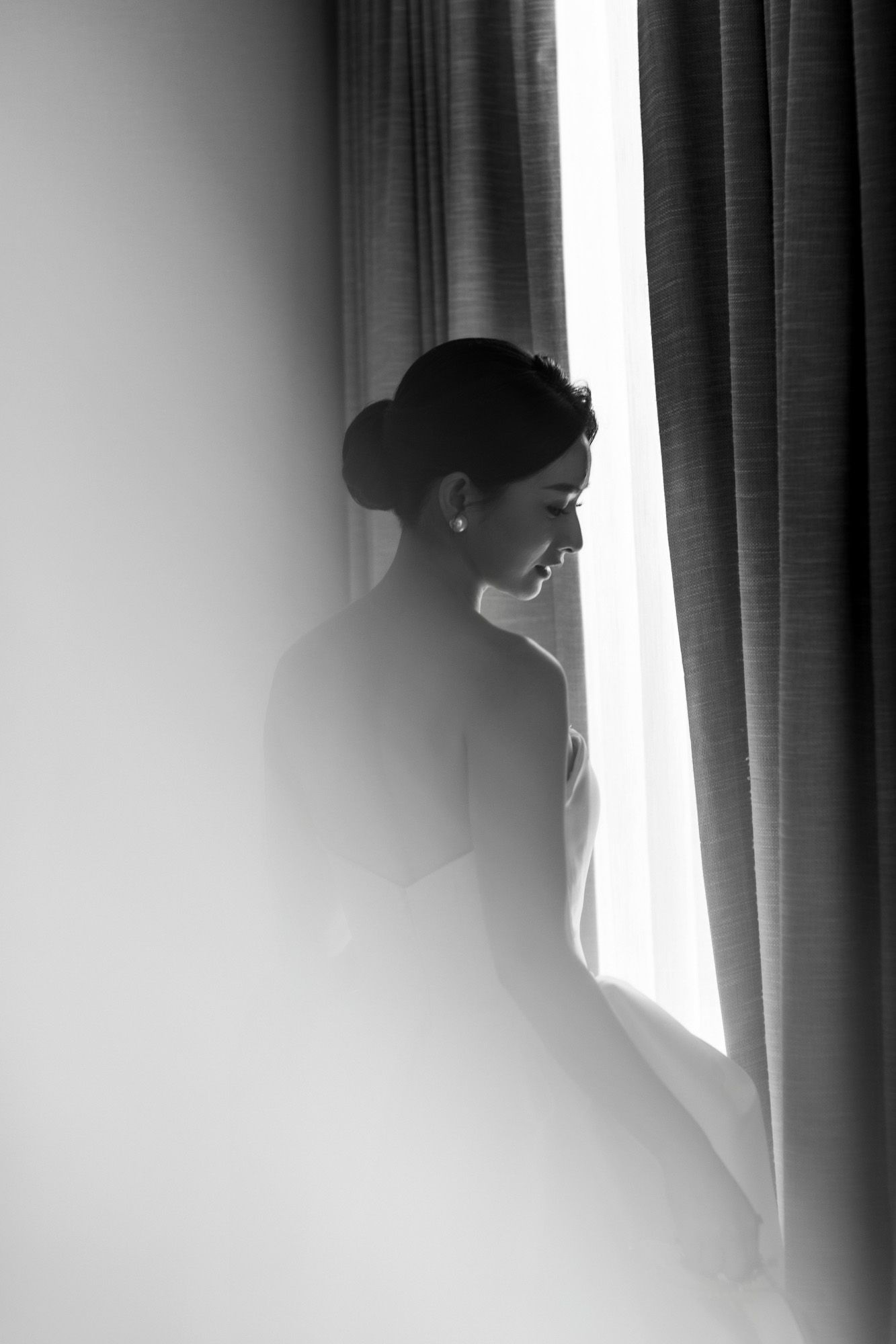 Silhouette of smiling bride in white wedding dress standing by curtained window, black and white.
