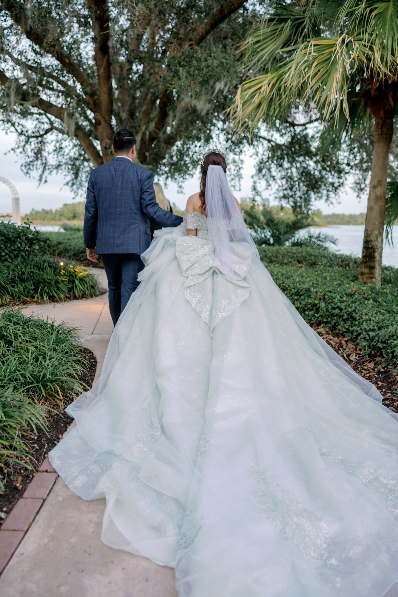 Bride in elegant white gown with long train walks with groom in blue suit along garden path near water.