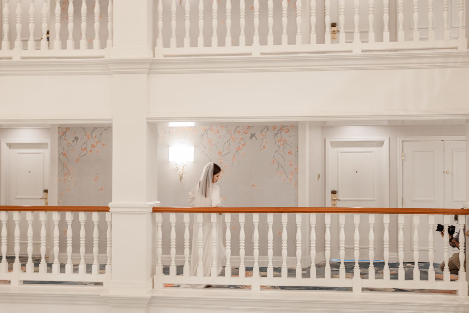 Bride in white gown standing on elegant hotel balcony with white railings and decorative wallpaper.