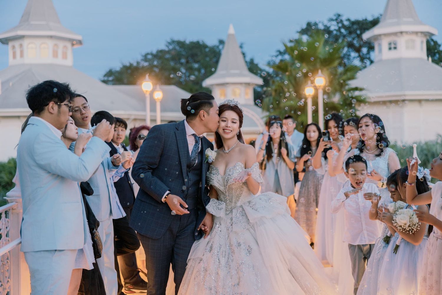 A couple shares a romantic kiss at their wedding reception while guests celebrate with sparklers at a fairy-tale venue.