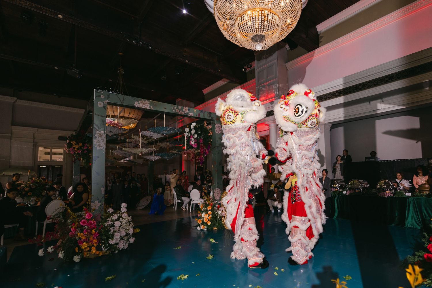 Two lion dancers in white and red costumes perform under a crystal chandelier at an elegant banquet hall celebration with floral decor.